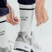 Person wearing white socks with 'Call Me Daddy' text and gray sneakers on a light background